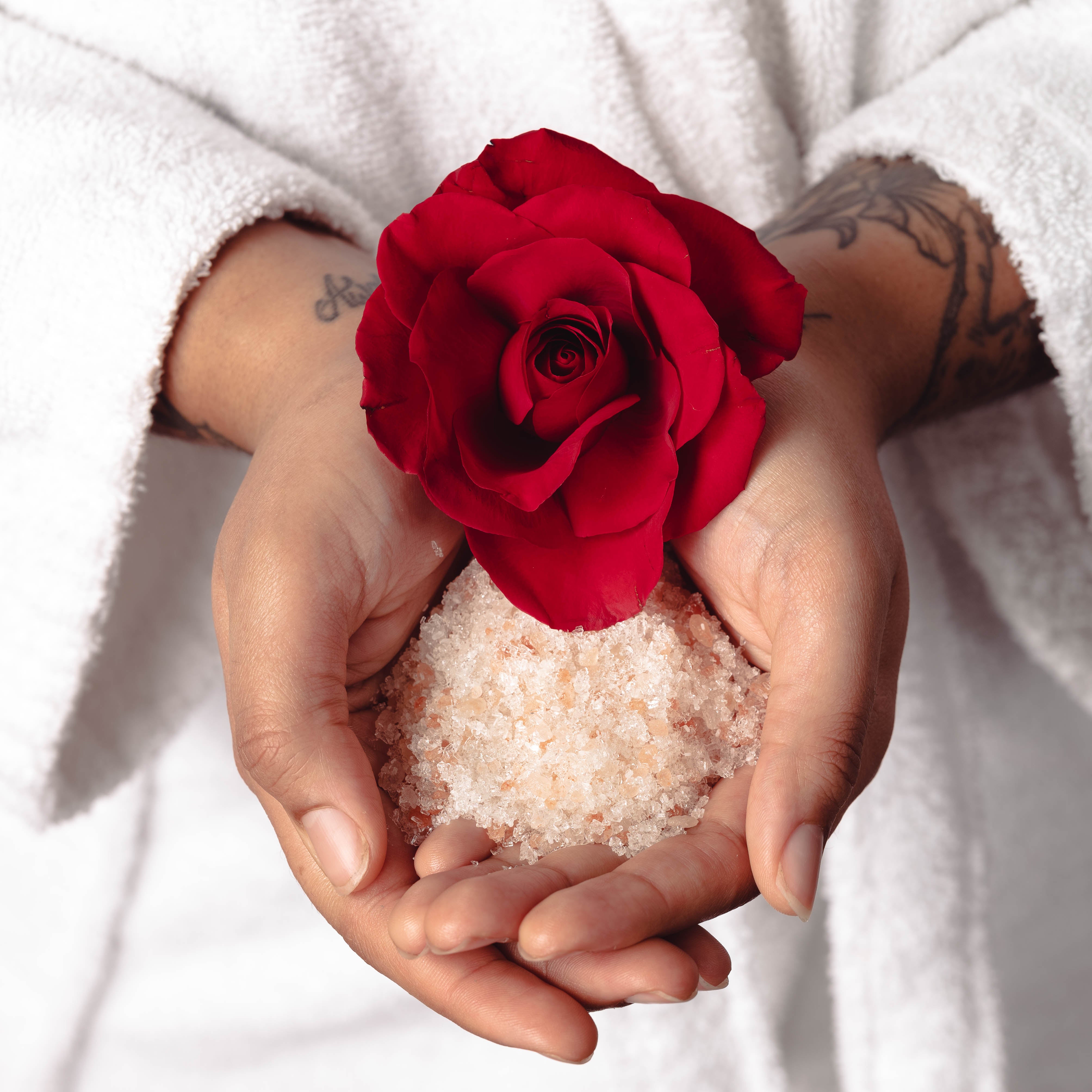 Two hands holding rose essential oil scented bath soaking salts with a red rose. 