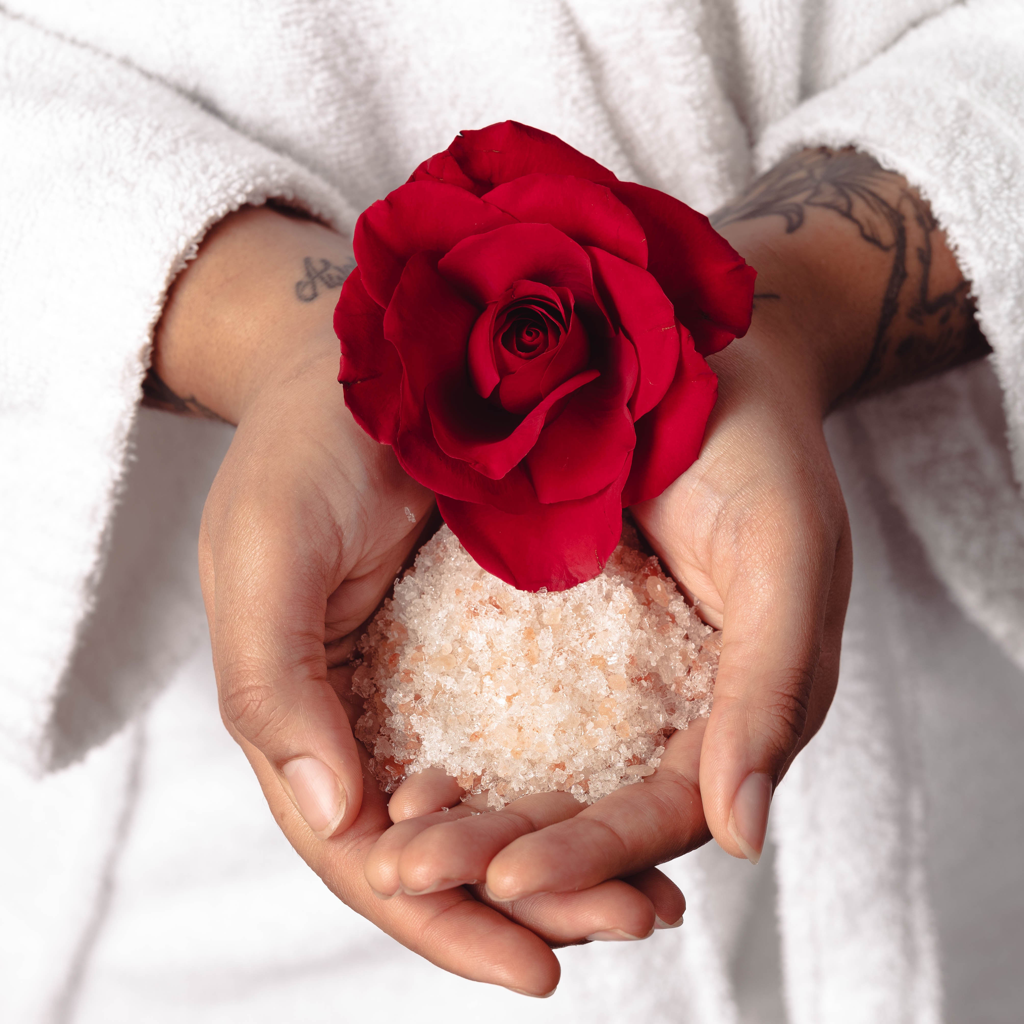 Two hands holding rose essential oil scented bath soaking salts with a red rose.