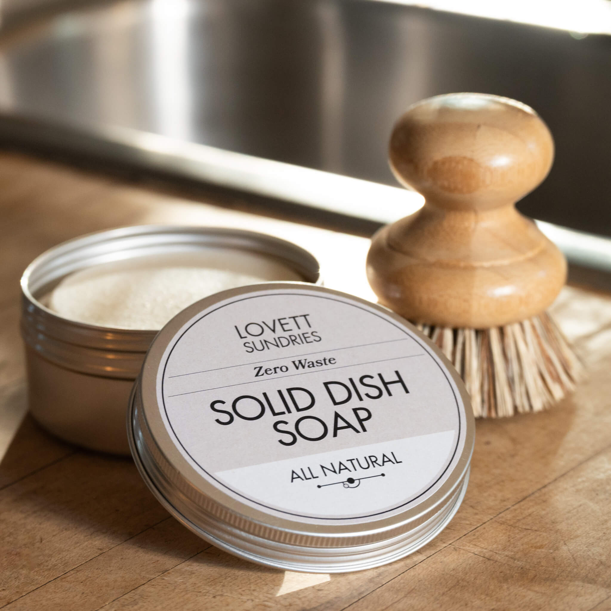 Solid Dish Soap in a tin with a brush on a wooden counter in the evening sun. 