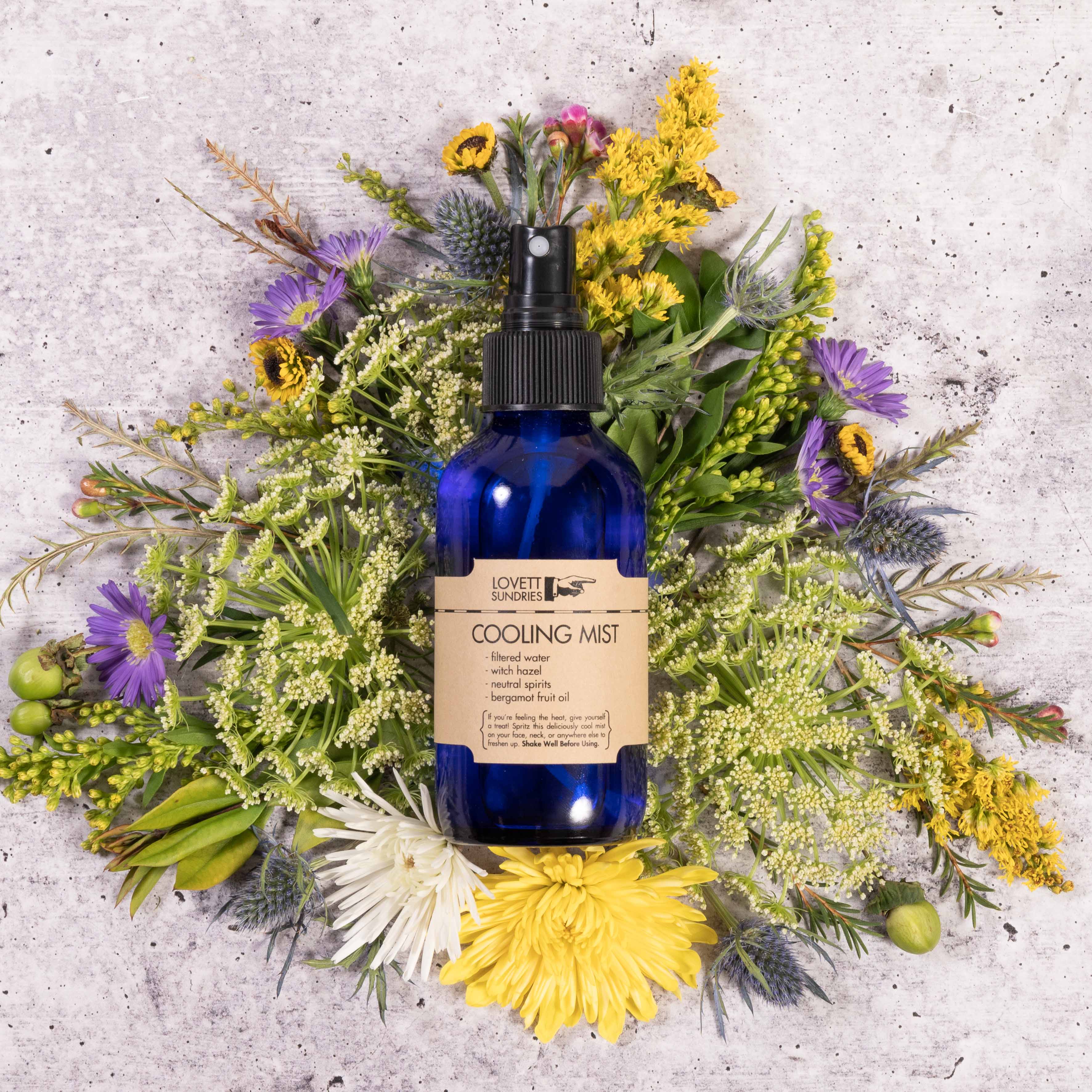 Blue bottle of cooling mist surrounded by vibrant wildflowers on concrete background.