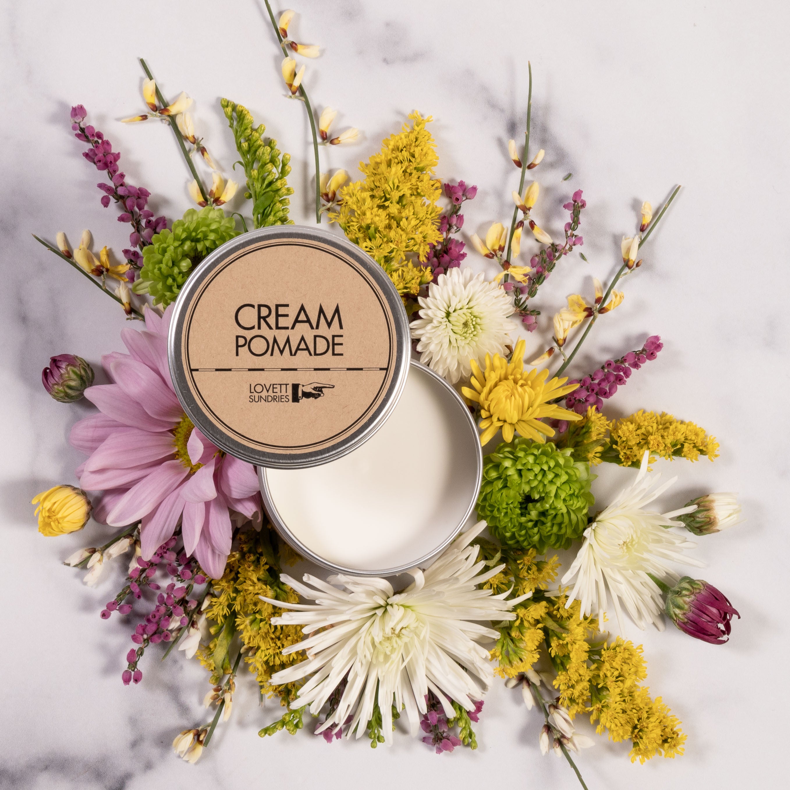 Cream pomade in a tin surround by colorful flowers on marble surface