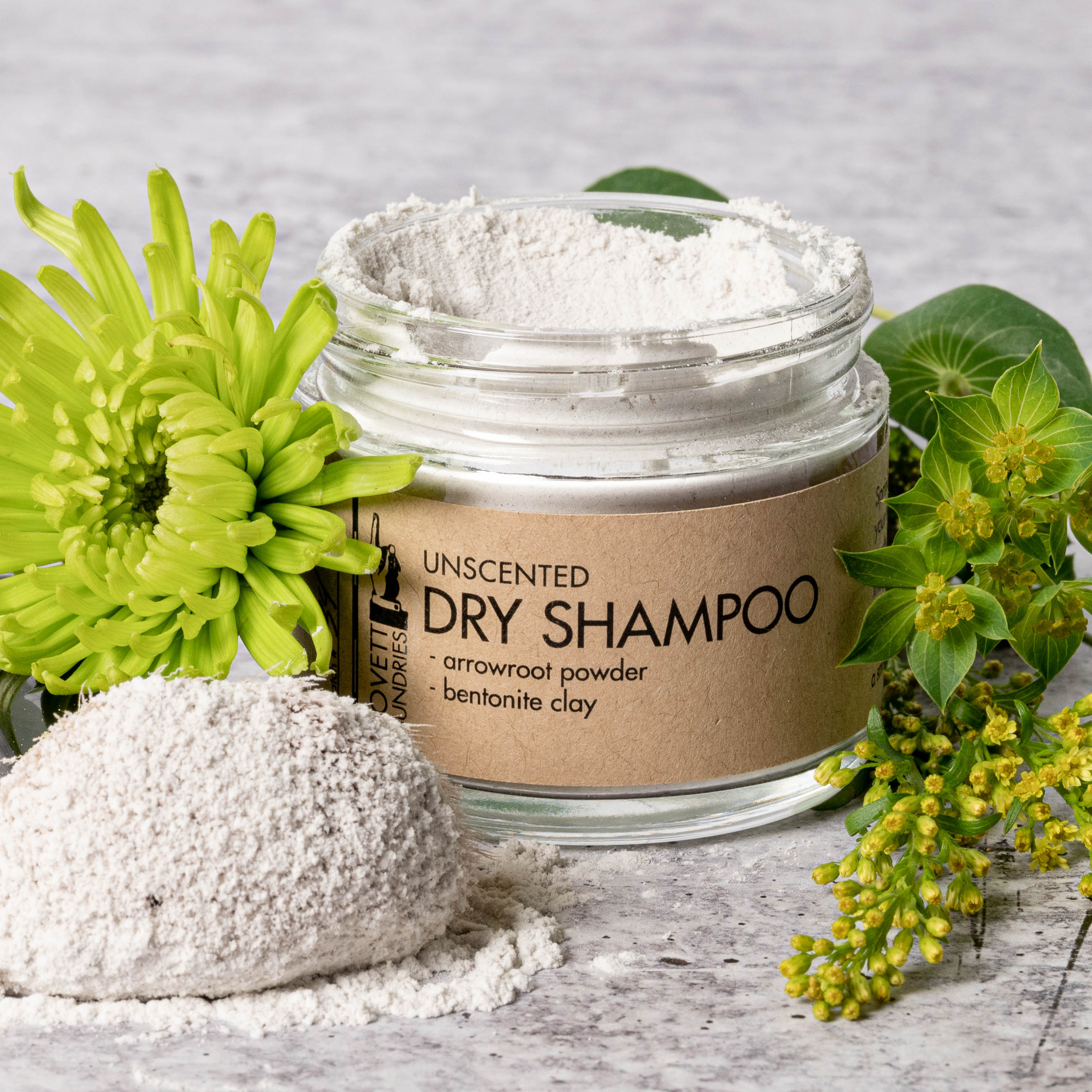 A glass jar of unscented natural dry shampoo surrounded by plants and clay