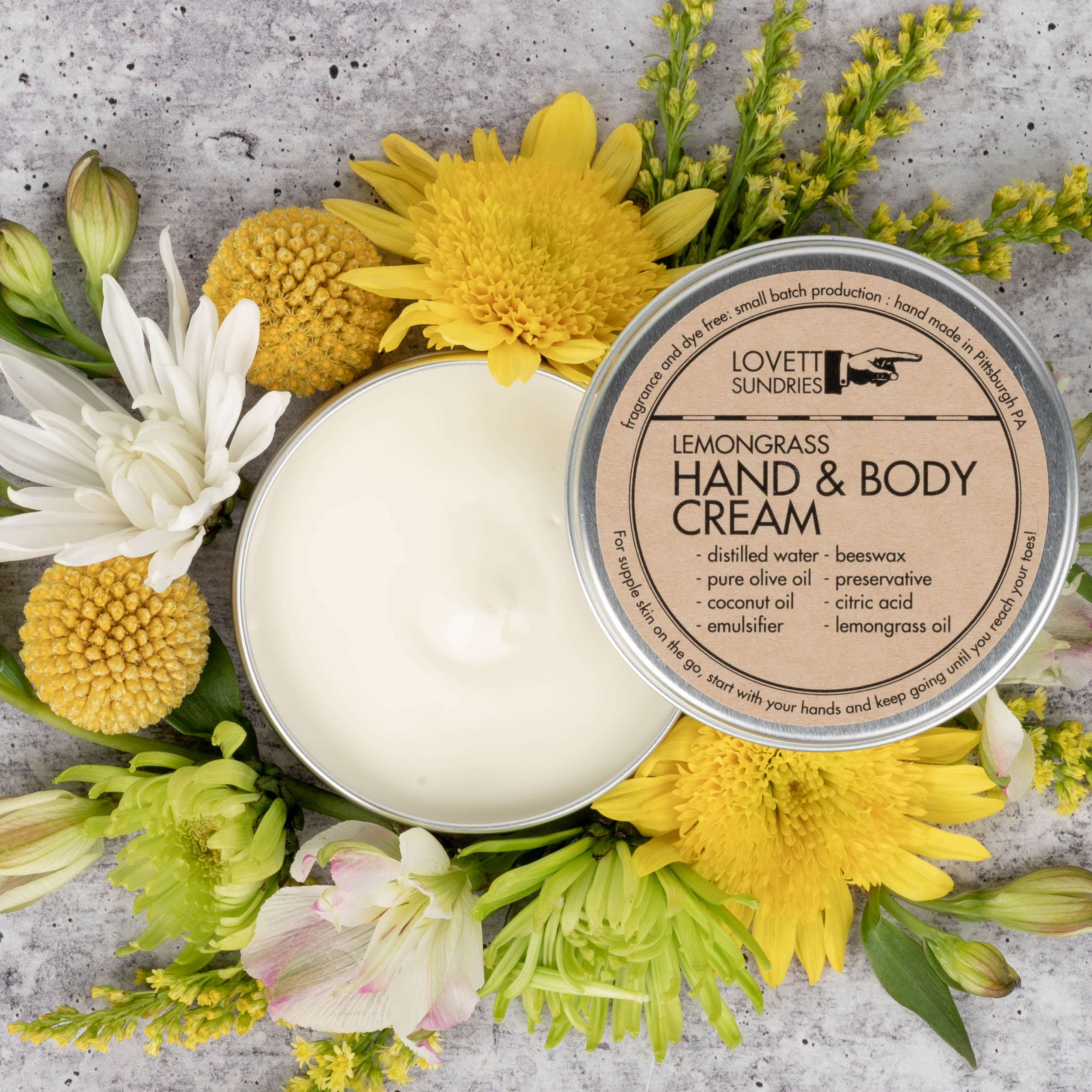 An open tin of lemongrass Hand & Body Cream surrounded by flowers.