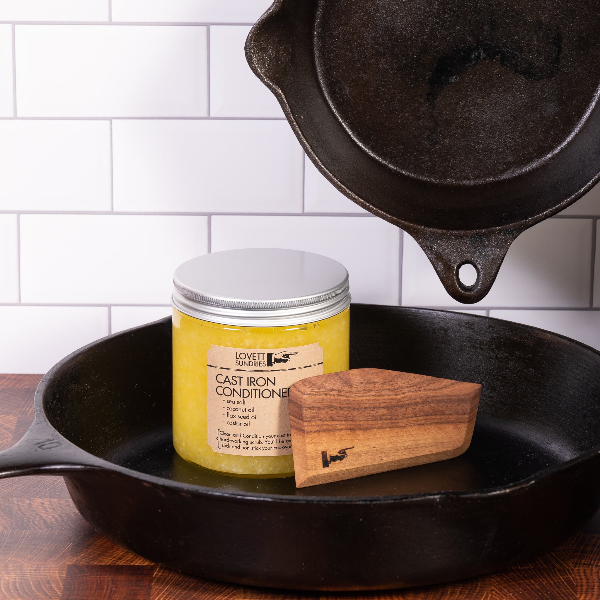 Hardwood cast iron scraper in pan with a jar of cast iron conditioner and skillets on a wooden kitchen counter. 